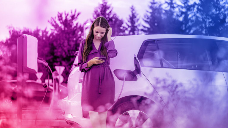 E-Auto lädt an einer Wallbox, eine Frau schaut dabei auf ihr Smartphone