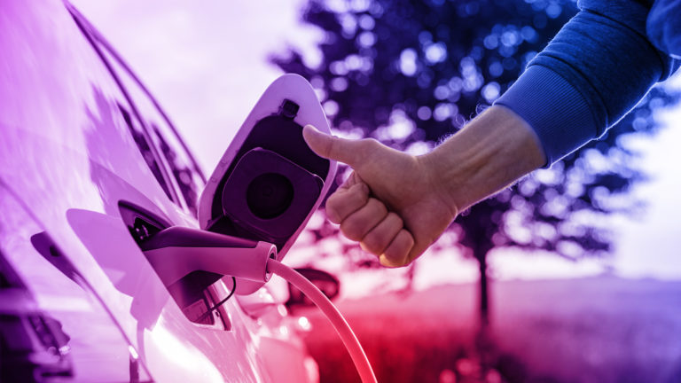 Elektroauto wird geladen in der Natur. Fahrer zeigt Daumen hoch.